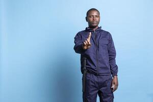 African american security officer raising a finger in front of the camera, showing a restriction symbol against blue background. Man working as a patrol agent presents a stop sign in studio. photo