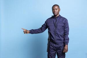 African american man in security agent uniform points to the left or right side, does a commercial advertisement against blue background. Bodyguard indicating a direction with his index finger. photo