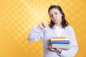 Portrait of woman with pouting expression on face holding pile of books, showing disproval of reading hobby. Sulky lady with stack of novels in arms doing thumbs down hand gesturing, studio background photo