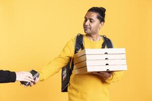 Smiling delivery worker offering takeaway order to customer paying with debit card. Client buying meal from courier, swiping card on POS device, isolated over studio background photo