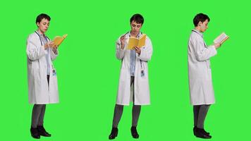 Medic specialist reading a healthcare literature book to develop her medical expertise in the industry, stands against greenscreen backdrop. Clever doctor in white coat enjoys lecture. Camera A. photo