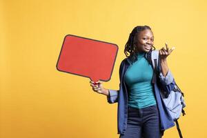 Jolly BIPOC girl holding red speech bubble sign of empty copy space for message. Smiling african american person presenting thought bubble cardboard used as promotion concept, studio background photo