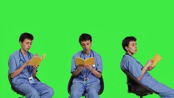 Health specialist is reading a literature book to acquire physician specialization and surgeon expertise, sitting in a chair. Nurse studying healthcare industry lecture against greenscreen. Camera B. photo