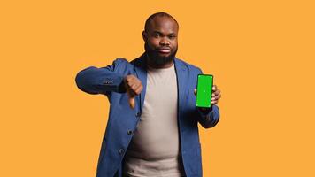 Upset african american man showing thumbs down sign gesturing holding green screen phone, disapproving. BIPOC person doing rejection hand gesture regarding smartphone, studio background, camera B photo