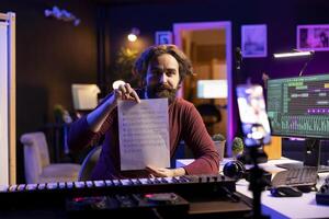 Talented artist explaining sheet with musical notes on smartphone vlog, doing online lessons in his home studio. Producer musician teaching people how to read music sheet, piano keyboard. photo