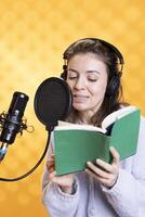 Smiling narrator wearing headphones reading aloud from book into microphone against yellow background. Cheerful person recording audiobook, creating engaging media content for listeners photo