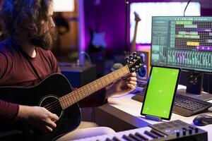 Young artist learning to tune his acoustic guitar with online tutorials via greenscreen tablet in home studio, playing musical instrument. Producer practicing strings accords and recording tunes. photo