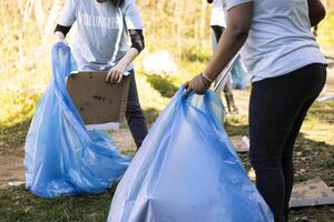 Women volunteers tidying the forest area of junk and plastic waste, collecting garbage with claw and blue disposal bag. Team of activists volunteering to preserve natural environment. photo
