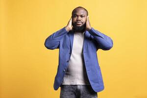 African american man troubled by excessive noise, suppressing it. BIPOC person covering ears with hands, annoyed by loud surrounding sounds, isolated over studio background photo
