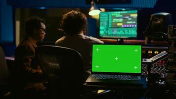 Sound designer and singer look at laptop with isolated display in control room, operating mixing console and volume level buttons to create a hit song. Artist working with audio expert. Camera B. photo