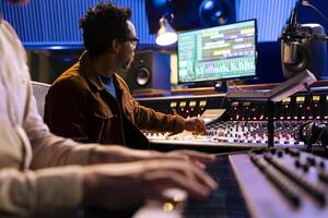 African american audio expert records artist playing piano in studio, using mixing console and digital editing software on pc. Producer composing new songs with musician on electronic keyboard. photo