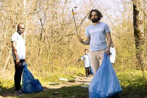 Activist using claw tool to grab rubbish and plastic waste from the ground, storing garbage in a trash bag and clean the natural environment. Male environmentalist gathering litter. photo