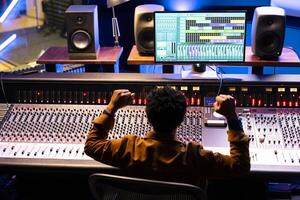 Confident sound engineer finishing a new hit song in professional music studio, working with digital software on pc and console in control room. African american producer creating records with gear. photo
