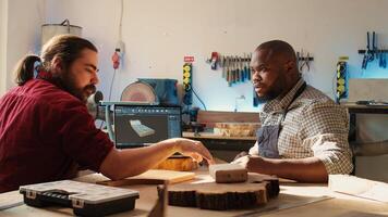 Happy BIPOC cabinetmaker and colleague comparing wood piece with schematic displayed on laptop. Cheerful carpenters in joinery crosschecking wooden object with notebook blueprint photo