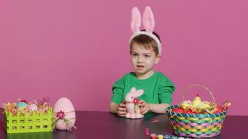 Adorable little child playing with a stuffed rabbit and a pink egg at table, having fun decorating festive easter ornaments in studio. Cheerful happy boy enjoying holiday preparations. Camera A. photo