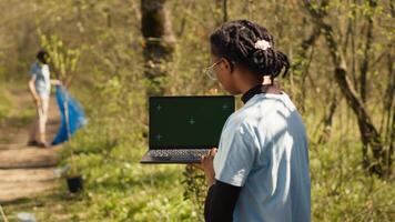 African american girl cleaning up forest and using isolated laptop display, taking part in a litter cleanup activity to protect the ecosystem and wildlife. Nature preservation concept. Camera A. photo