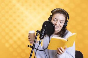 Smiling woman reads ebook from ereader, records audiobook using microphone, drinking coffee, studio background. Jolly narrator using mic to record novel, reading text from tablet, holding beverage cup photo