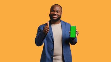 Happy african american man holding green screen phone, doing recommendation. Cheerful BIPOC person presenting mockup device, giving thumbs up sign, studio background, camera B photo