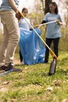 Environmentalists picking up trash from the natural forest environment by grabbing plastic and junk objects from the forest, collecting in a garbage bag. Using tools to pick up rubbish. photo