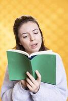 Shocked woman browsing through intriguing book pages, gasping, having fun, isolated over studio background. Surprised person reading stunning novel with plot leaving her with mouth open photo