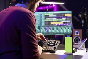 Music producer uses controls and knobs on mixing console in home studio, having a smartphone with greenscreen display. Artist learning to edit tunes and add sound effects with daw software. photo