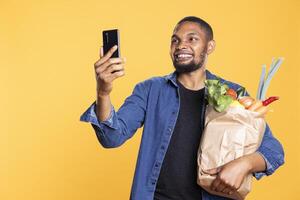Charismatic relaxed person taking a selfie with his bag of bio groceries, enjoying a successful eco friendly shopping session. Happy confident guy takes photos on his smartphone app.