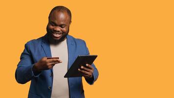 Smiling man scrolling on tablet touchscreen, using internet and credit card to pay for products. African american person happily browsing online shopping websites, studio background, camera A photo