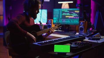 Songwriter playing acoustic guitar in home studio with greenscreen display running on smartphone app. Musician practices singing on musical instrument with strings, isolated screen. Camera B. photo