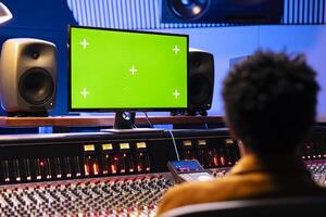 African american producer looks at greenscreen on monitor and mixing music with control desk panel board. Audio expert working in professional studio to adjust volume and add sound effects. photo
