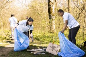 Diverse volunteers working to clear the forest from trash, fighting pollution to protect the natural ecosystem. Young team of activists collecting junk and plastic waste with tools. photo