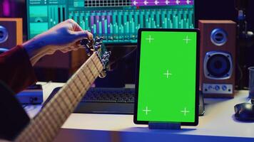 Audio engineer practices tuning his acoustic guitar in home studio, watching a tutorial online via tablet with greenscreen. Musician learning to play instrument and create music. Camera A. photo