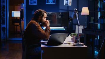 Technician focused on typing complex code in neon lit home office, developing software application. It specialist removing headphones to better concentrate on difficult task, camera B photo