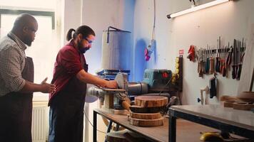 Man teaching african american trainee how to shape wooden bowl on disc sander in assembling shop. Worker learning from woodworking professional how to use grinding machine rotating disc, camera B photo
