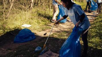 Environmental activist picking up trash with a claw tool and recycling, protecting the natural forest habitat and doing litter cleanup. Woman doing voluntary work to stop pollution. Camera B. photo