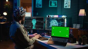 IT admin coding and using EEG headset to upload brain into green screen laptop, gaining immortality. Neuroscientist engineering transhumanism experiment on chroma key device, camera B photo