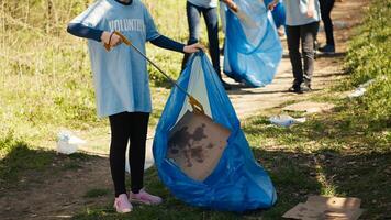 Small girl collecting rubbish around a forest with a claw tool and garbage bags, cleaning up the environment by recycling plastic waste and trash. Child learning to protect ecosystem. Camera A. photo