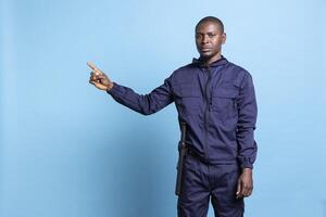 African american serious security agent in uniform pointing to the left side, creates a commercial ad to present something next to him. Guard raising his index finger, indicating a direction. photo