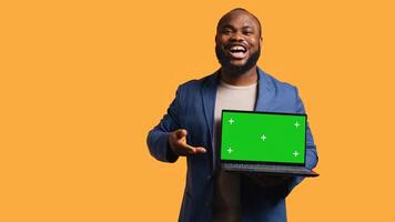 Smiling man holding isolated screen laptop, doing recommendation, talking with viewers. Cheerful BIPOC person pointing towards chroma key notebook, studio background, camera B photo
