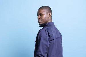 Portrait of african american security agent posing in front of camera, looking over his shoulder against blue background. Male bodyguard dressed in uniform, detecting any suspicious behavior. photo