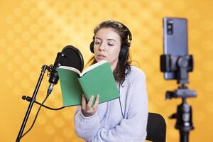 Influencer filming herself reading book with smartphone on tripod, creating audiobook, studio background. Woman doing vlog with mobile phone while producing digital recording on novel using microphone photo