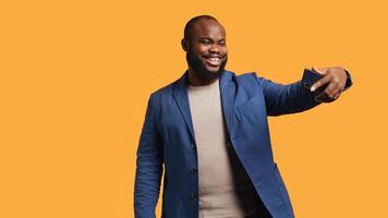 Joyous man using smartphone to take selfies and post them on social media. Happy BIPOC person taking photos using phone selfie camera, isolated over yellow studio background, camera B