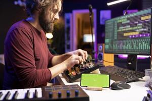 Audio engineer uses knobs on stereo gear for mixing and mastering session, having a smartphone with greenscreen display in home studio. Musician learning to edit tunes with daw software. photo