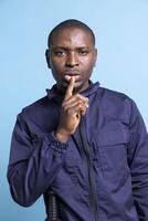 African american security officer putting a finger over lips for quietness, showing a hush sign against blue background. Man in safeguarding uniform does a mute symbol for silence in studio. photo