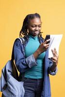 BIPOC young girl taking photos of school homework to send to colleague, isolated over studio background. Student photographing papers with information needed for university courses using phone