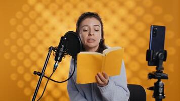 Influencer filming reading book with smartphone on tripod, creating audiobook, studio background. Woman vlogging with mobile phone while producing digital recording on novel using microphone, camera B photo