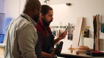 Cabinetmaker teaching apprentice to drill hole into timber block in studio. Woodworking pupil learning from master in joinery how to insert nail into piece of wood using power drill, camera A photo