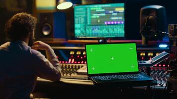 Audio technician leveling sound on recorder mixing console, having a laptop that shows greenscreen display. Producer turns up the volume of tracks in professional studio, control room setup. Camera A. photo