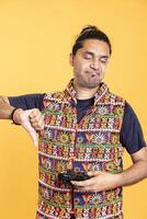 Portrait of annoyed man showing thumbs down sign holding gamepad, disapproving of videogames. Disgusted person protesting against gaming, doing rejection hand gesture, isolated over studio backdrop photo