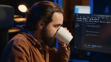 Focused programmer drinking coffee in apartment while solving database errors on computer. Remotely working IT admin concentrated on finalizing coding project in home office, camera A photo