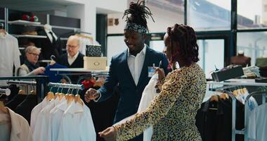 Retail assistant showing new clothing collection to customer, recommending colors and sizes for pregnant woman in department store. African american client asking worker for assistance. Camera B. photo